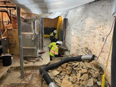 Staff in a basement with a heating oil leak. Air barriers and ventilation have been put in place, and the floor has been removed to reach the leak and contaminated soil. In the foreground, you can see a tube that leads back to the vacuum truck for debris removal.